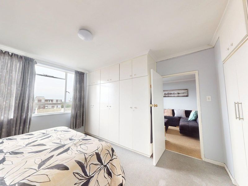 A bedroom featuring extensive built-in cupboards, neutral carpeting, and a window providing natural light, with an adjacent view into the living area.