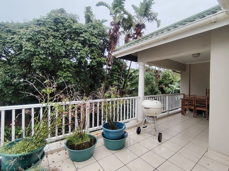 A covered balcony featuring tiled flooring and a white balustrade railing, overlooking a lush garden area.