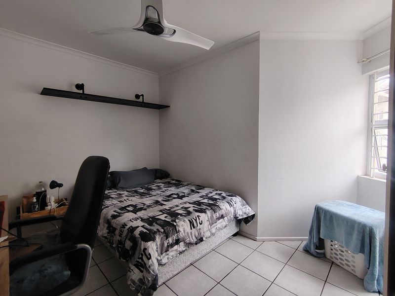 A functional bedroom featuring tiled flooring, a ceiling fan, and a wall-mounted shelf, offering a neutral space ready for personalization.