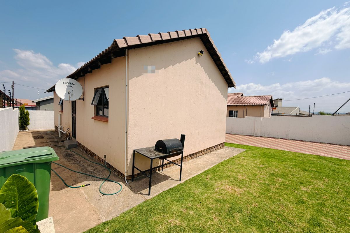 Rear view of the property featuring a tiled roof, paved driveway, and a small lawn area enclosed by boundary walls.