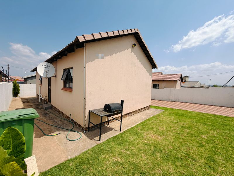 Rear view of the property featuring a tiled roof, paved driveway, and a small lawn area enclosed by boundary walls.