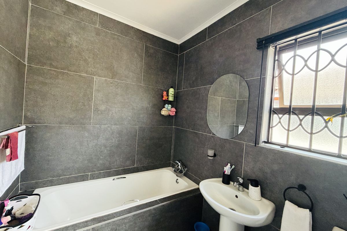 A modern bathroom featuring dark grey wall tiles, a built-in bathtub, and a white pedestal basin with a window fitted with security bars.