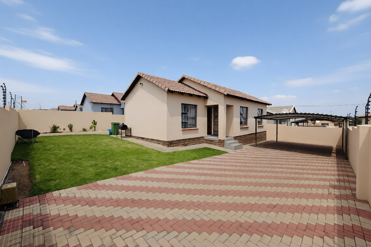 A well-maintained single-story house featuring a paved driveway, carport, neat lawn, and secure perimeter wall with electric fencing.
