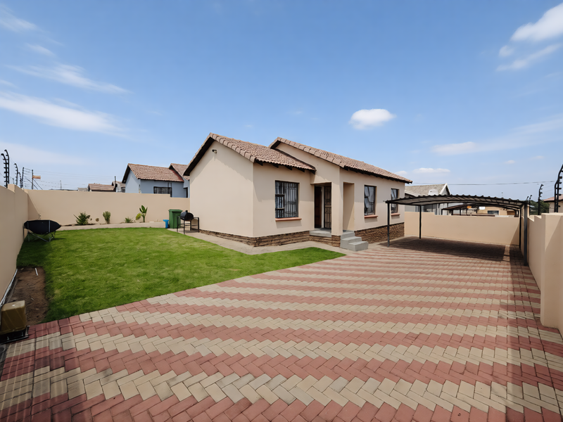 A well-maintained single-story house featuring a paved driveway, carport, neat lawn, and secure perimeter wall with electric fencing.