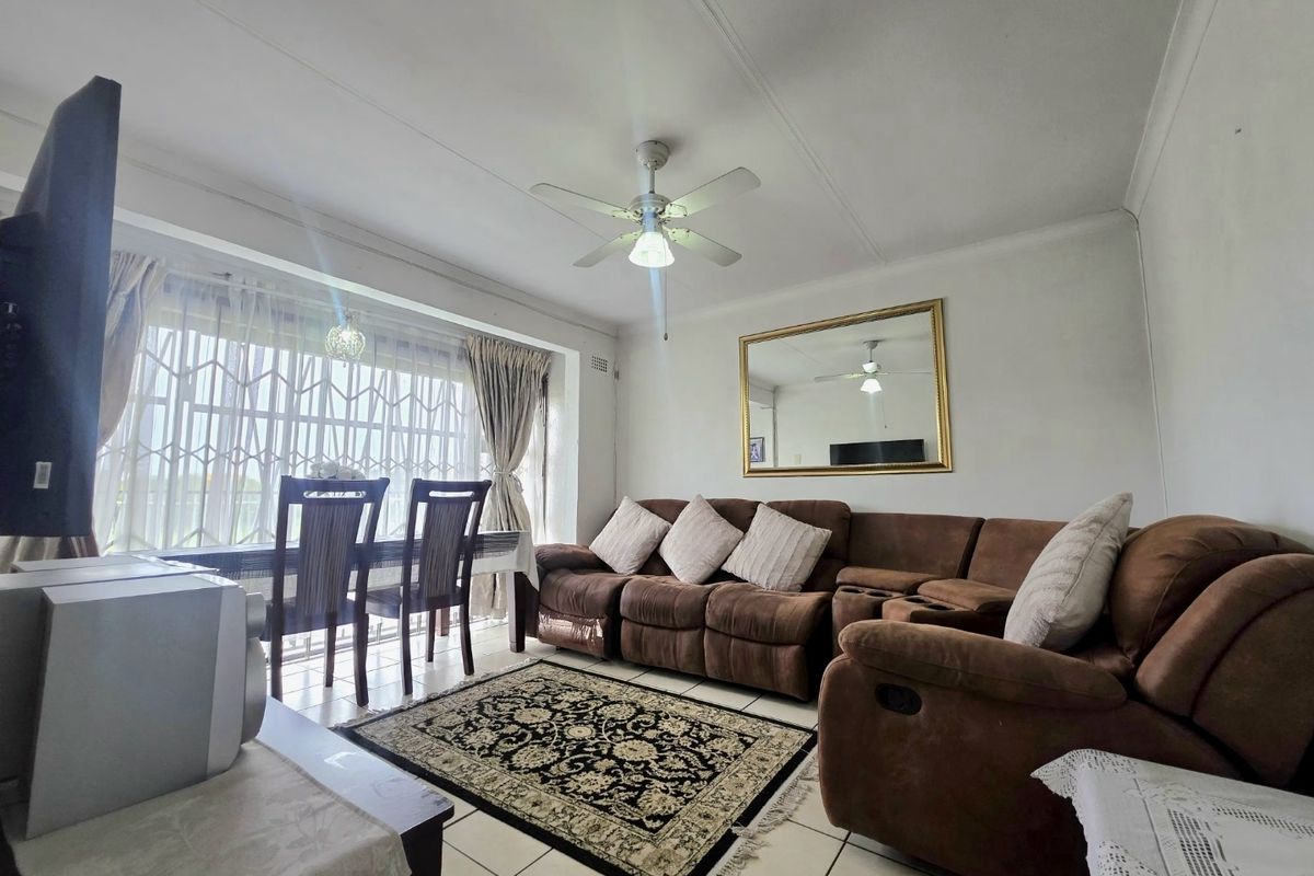 A spacious living room featuring tiled flooring, a ceiling fan, and a large window with security gate, offering a functional and comfortable living space.