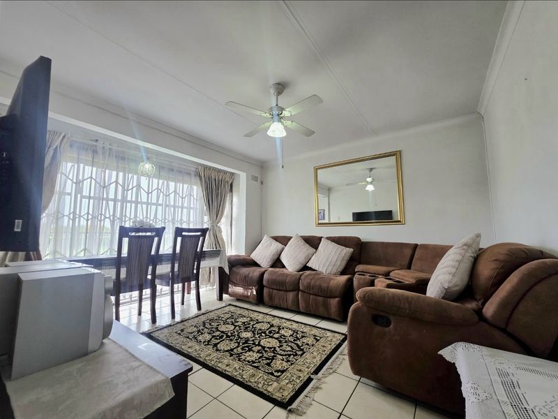 A spacious living room featuring tiled flooring, a ceiling fan, and a large window with security gate, offering a functional and comfortable living space.