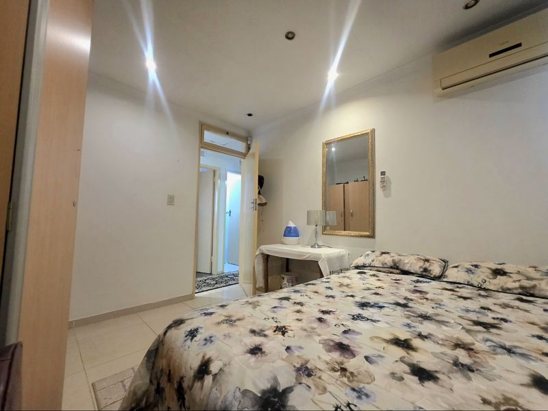 A functional bedroom featuring tiled flooring, recessed downlighting, a wall-mounted air conditioning unit, and built-in wardrobe storage.