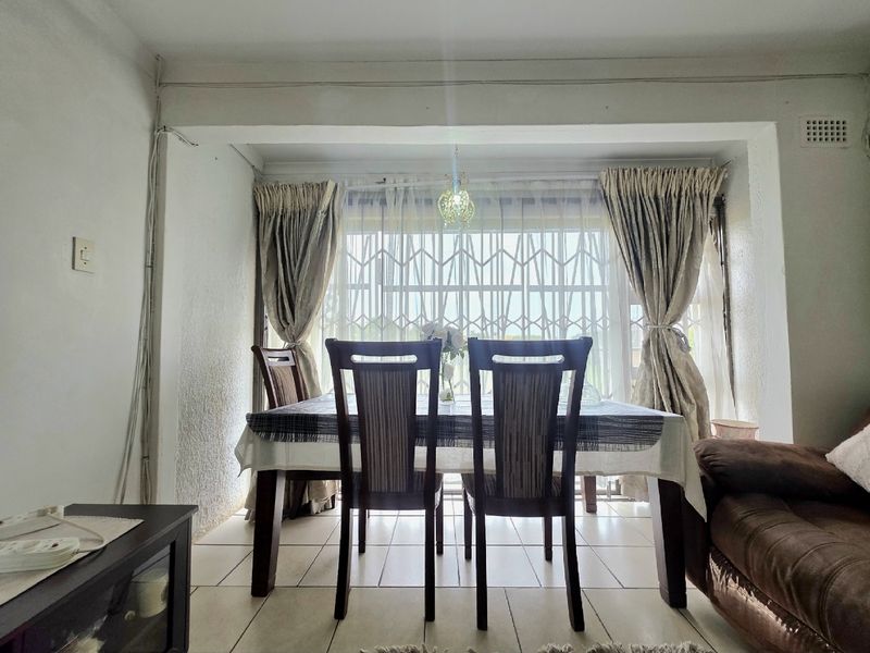 A dining area featuring tiled flooring, a window with a security gate, and a central light fixture, showing signs of dated electrical finishes.
