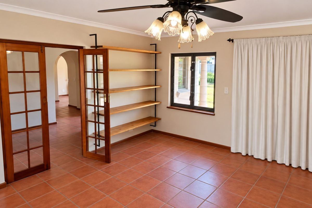 A bright, empty living room featuring terracotta floor tiles, built-in wooden shelving, a ceiling fan, and classic wooden french doors leading to an adjacent space.