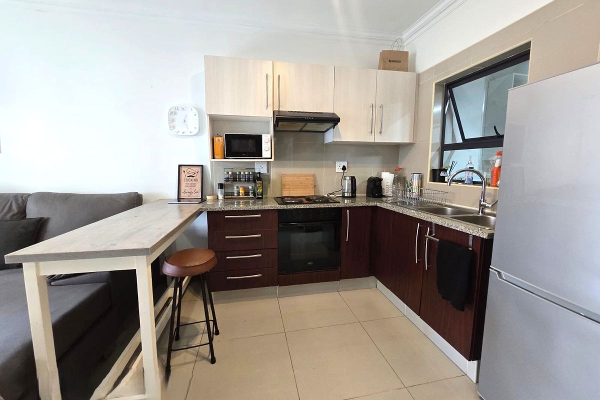 Compact open-plan kitchen featuring two-tone cabinetry, granite countertops, integrated oven and hob, and tiled flooring, seamlessly connected to the living area.