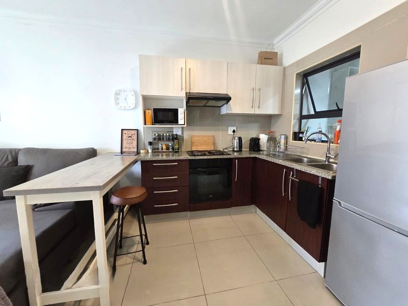 Compact open-plan kitchen featuring two-tone cabinetry, granite countertops, integrated oven and hob, and tiled flooring, seamlessly connected to the living area.