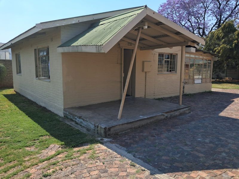Rear view of a single-story brick house featuring a covered concrete patio, corrugated metal roof, and paved outdoor area.