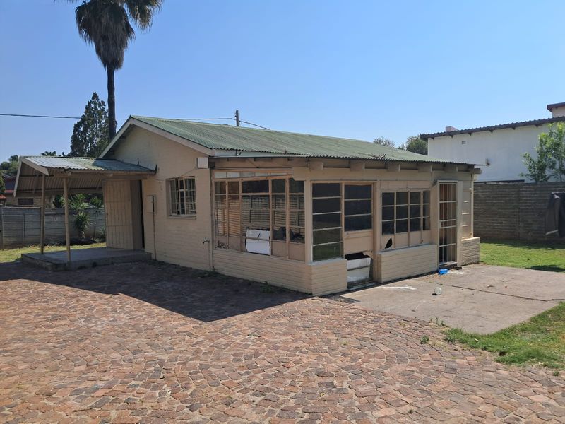 Rear view of a detached dwelling featuring a corrugated metal roof, brick-paved driveway, and a concrete patio area with visible maintenance requirements.