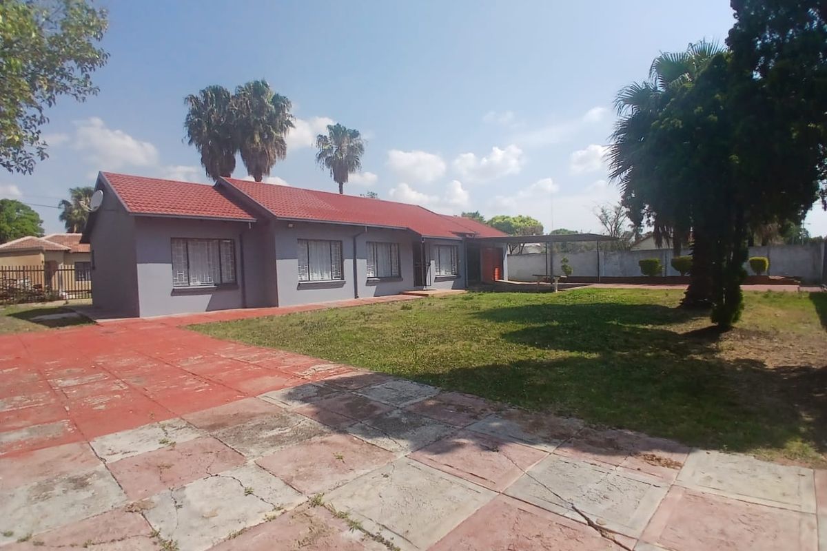 Rear view of a single-story house featuring a tiled roof, paved driveway, and a covered patio area set within a spacious garden.