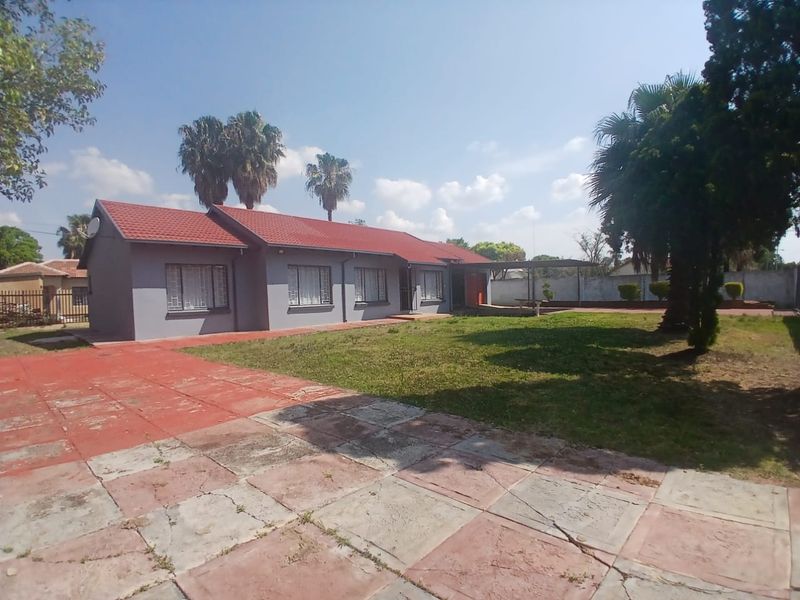 Rear view of a single-story house featuring a tiled roof, paved driveway, and a covered patio area set within a spacious garden.