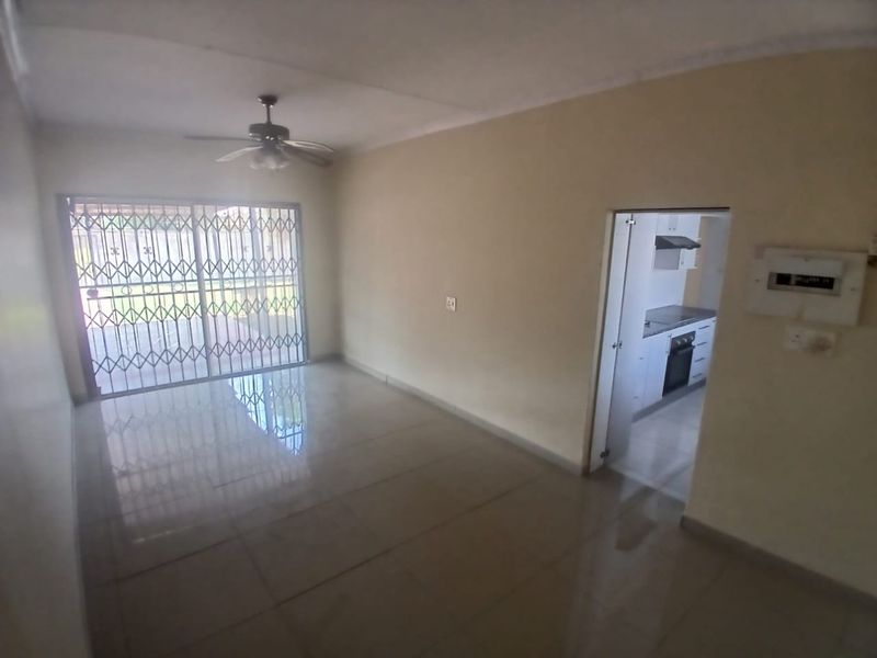 An empty living room featuring tiled flooring, a ceiling fan, and a security gate leading to the exterior, with an adjacent kitchen view.