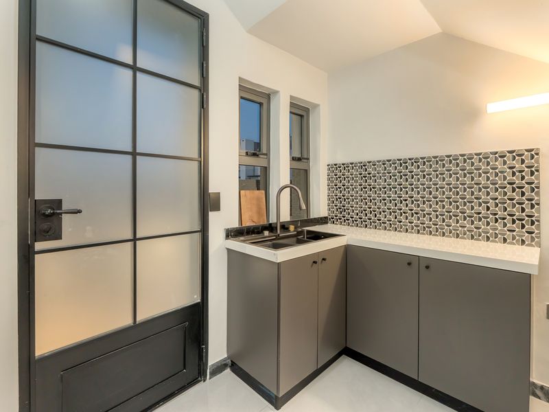 A modern, well-appointed scullery featuring sleek grey cabinetry, stone countertops, a geometric tile backsplash, and a stylish black-framed glass door.