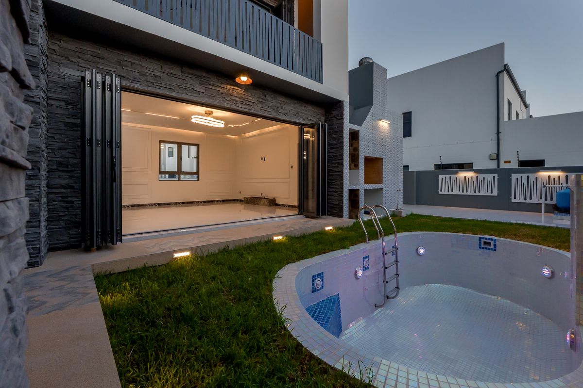 A modern rear exterior featuring a swimming pool, built-in braai area, and large stacking doors opening into a spacious, well-lit interior living area.