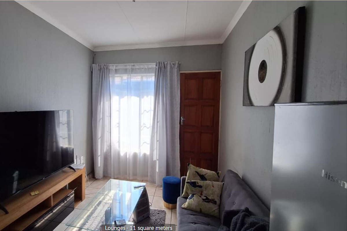 A compact living room featuring tiled flooring, a timber entrance door, and natural light through a window with curtain rails.