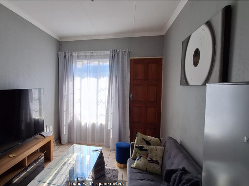 A compact living room featuring tiled flooring, a timber entrance door, and natural light through a window with curtain rails.