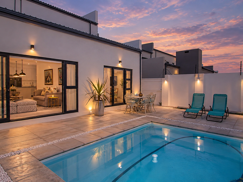 A beautifully maintained rear patio area featuring a swimming pool, paved terrace, and seamless indoor-outdoor flow through large sliding glass doors at sunset.