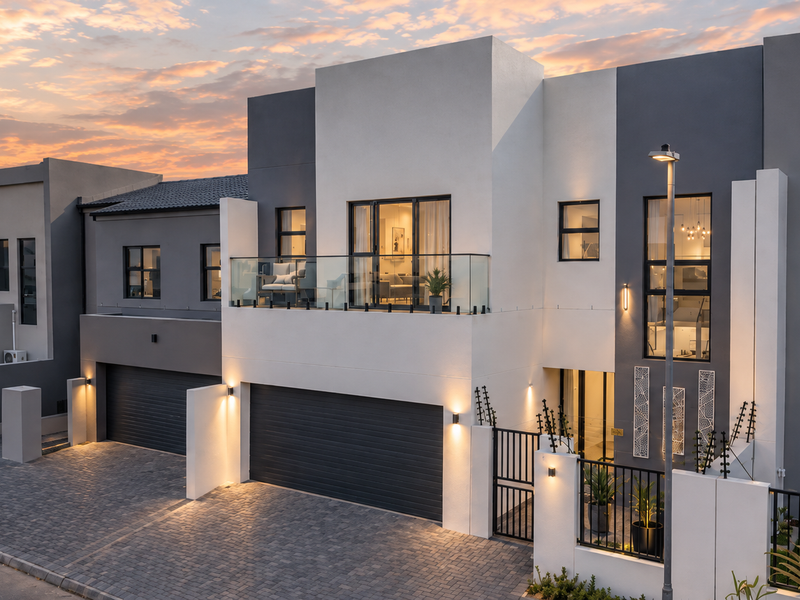 Modern double-storey townhouse facade featuring a double garage, paved driveway, and a private balcony with glass balustrades at sunset.