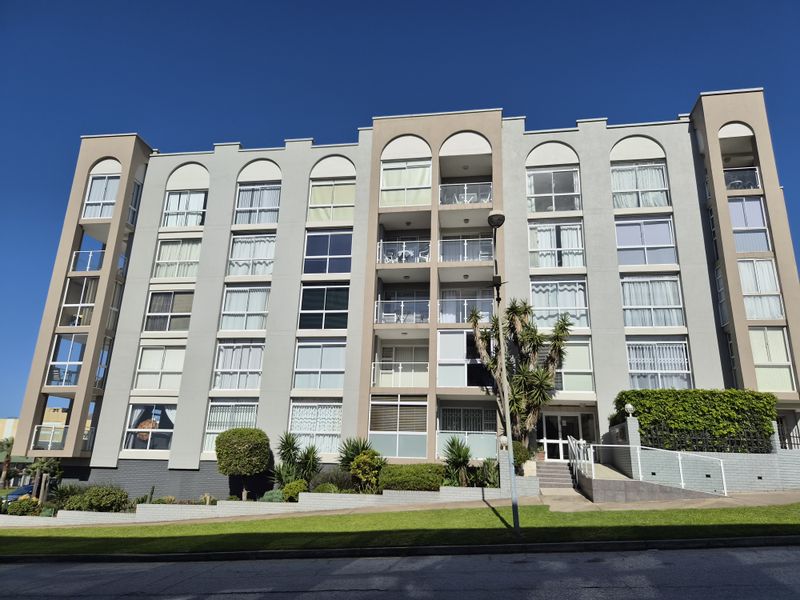 A clear front elevation view of a multi-story apartment building featuring balconies, aluminum windows, and a landscaped entrance area.