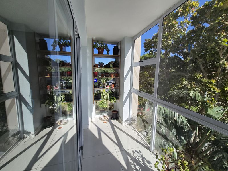 A bright, enclosed balcony featuring tiled flooring and large aluminium-framed windows, offering a pleasant outlook with natural light.