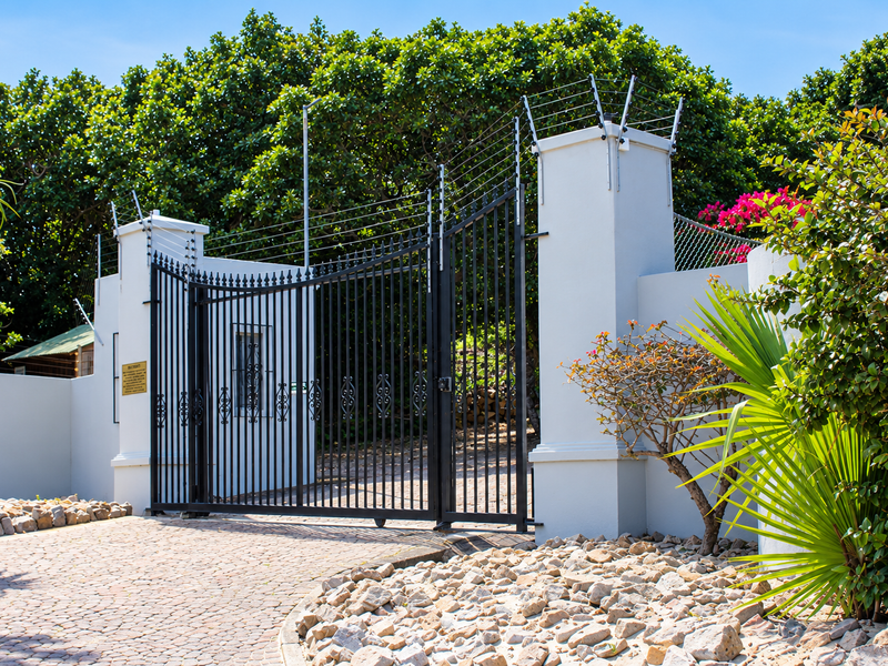 A secure residential entrance featuring a black wrought iron gate, masonry pillars with electric fencing, and a well-maintained cobblestone driveway.