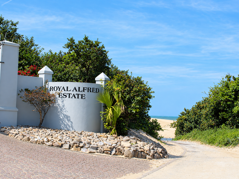 The entrance to Royal Alfred Estate featuring a secure gate, masonry wall, and paved driveway leading towards the coastal landscape.