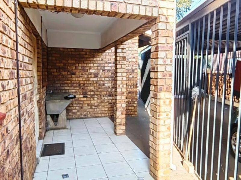 A covered outdoor patio area featuring face brick walls, tiled flooring, and a built-in utility sink, adjacent to a secure gated parking area.