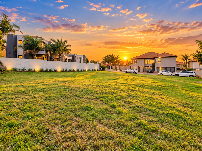 A vacant residential plot featuring a well-maintained grass lawn, retaining wall, and integrated landscape lighting, set within a developed estate at sunset.