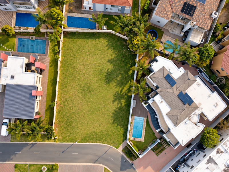 An aerial view of a vacant residential plot situated between established homes, featuring a clear lawn area and direct access to the paved street.