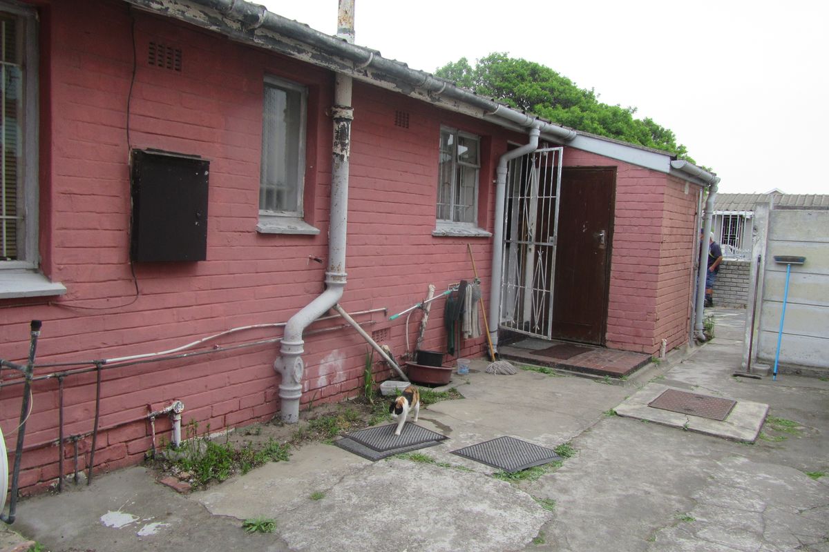 Rear exterior view of the property featuring painted brick walls, concrete paving, and external plumbing infrastructure requiring maintenance.