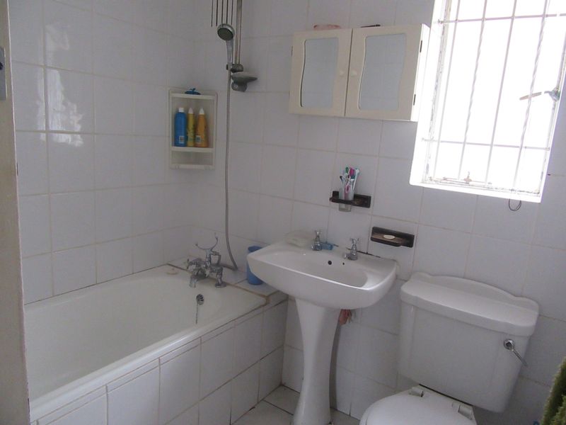 A functional bathroom featuring a pedestal basin, bathtub with shower mixer, and toilet, finished with white wall tiles and security bars on the window.