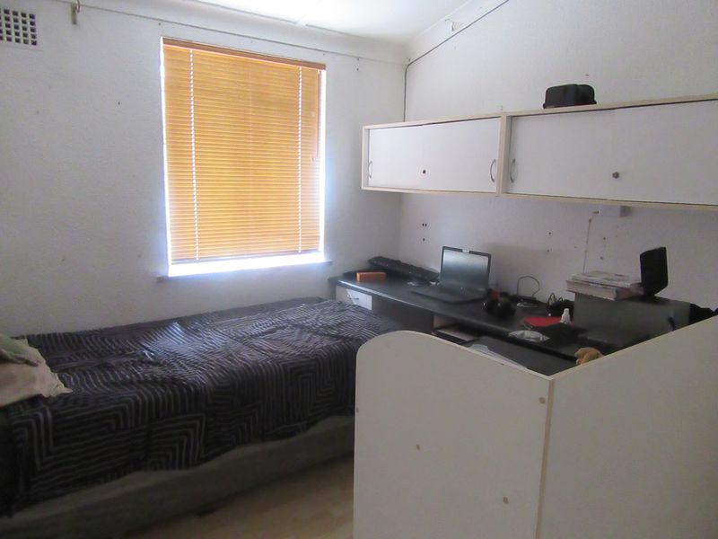 A compact bedroom featuring built-in desk space and overhead cabinetry, with a window fitted with venetian blinds.