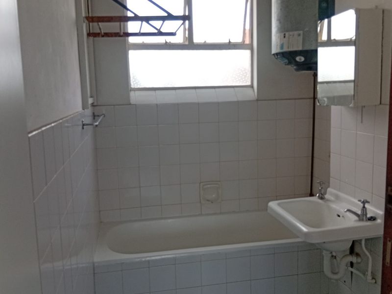 A functional bathroom featuring a bathtub, wall-mounted basin, and tiled walls. The space includes an exposed water heater and requires cosmetic updates.