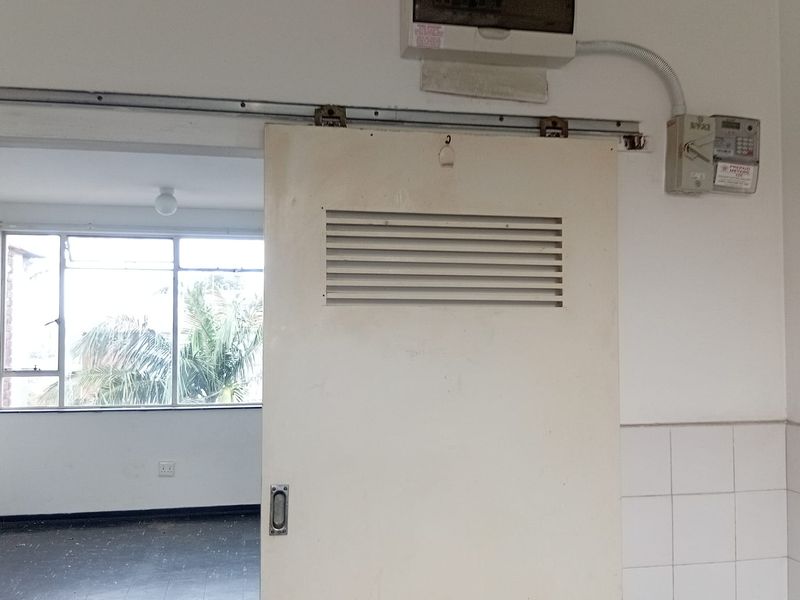 Utility area featuring a sliding door, electrical distribution board, and prepaid meter, leading into an adjacent room with large windows.