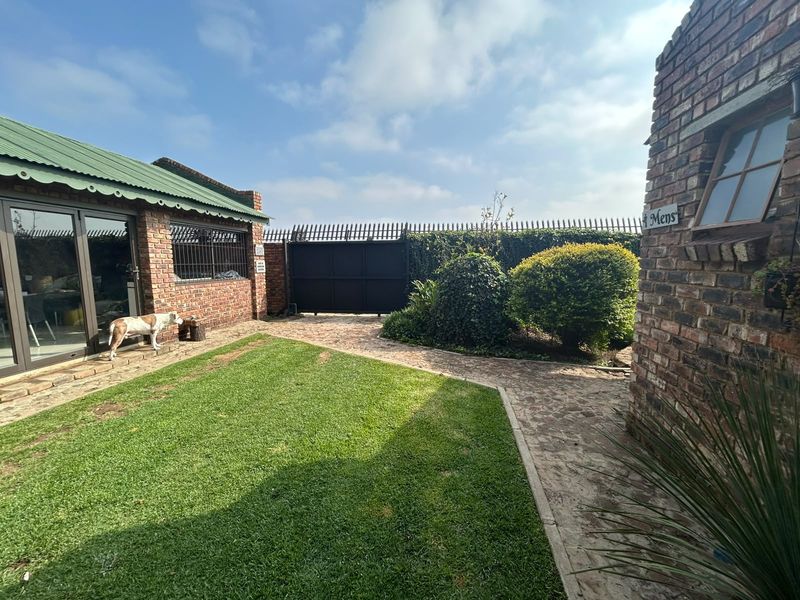 A well-maintained rear courtyard featuring face brick structures, a green corrugated roof, paved pathways, and a manicured lawn area.