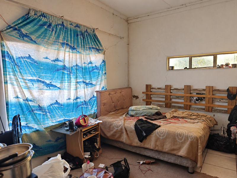 A bedroom featuring tiled flooring and a horizontal window, currently showing significant signs of wear including cracked wall plaster and peeling paint.