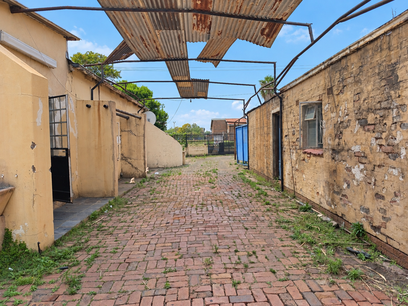 Rear exterior area featuring brick paving and a damaged metal roof structure, showing significant signs of weathering and structural deterioration.