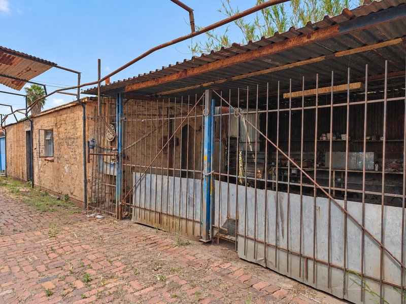 An outdoor utility or workshop area featuring a corrugated iron roof, steel security gating, and brick paving, showing significant signs of wear and weathering.