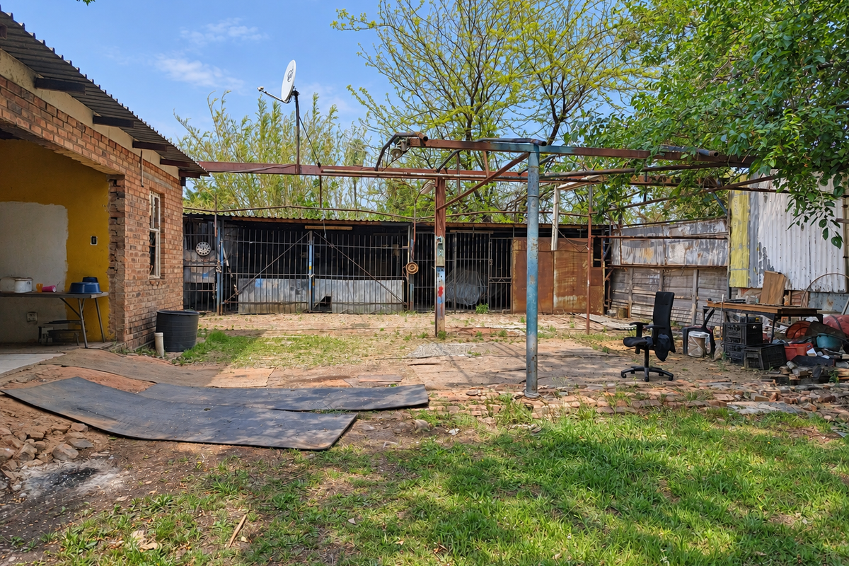Rear yard area featuring a brick facade, rusted metal overhead structure, and perimeter metal fencing, offering potential for landscaping or outdoor renovation.