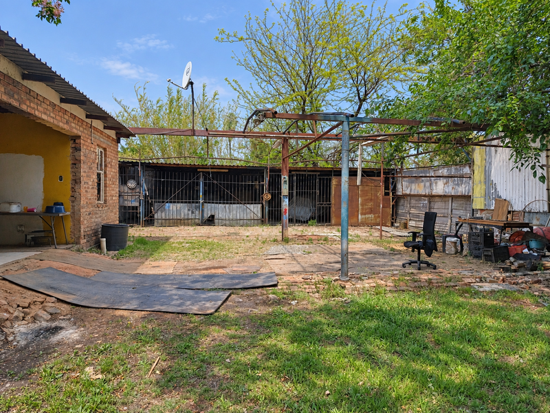 Rear yard area featuring a brick facade, rusted metal overhead structure, and perimeter metal fencing, offering potential for landscaping or outdoor renovation.
