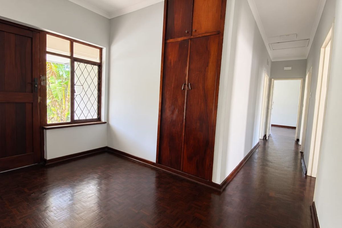 A bright, empty bedroom featuring polished parquet flooring, built-in wooden cabinetry, and a classic diamond-pattern window, leading into a connecting hallway.