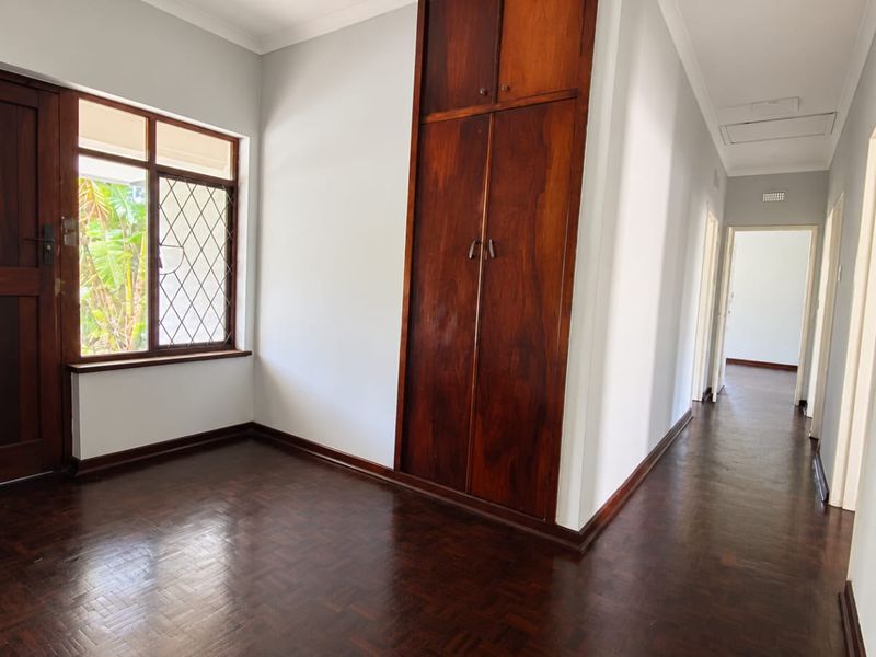 A bright, empty bedroom featuring polished parquet flooring, built-in wooden cabinetry, and a classic diamond-pattern window, leading into a connecting hallway.