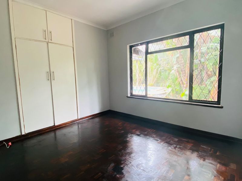 A bright, empty bedroom featuring polished parquet flooring, built-in storage cupboards, and a large window with security bars overlooking the garden.