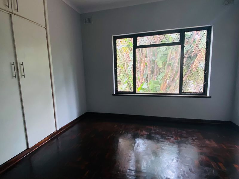 A bright, empty bedroom featuring polished parquet flooring, built-in cupboards, and a large window with security bars overlooking the garden.