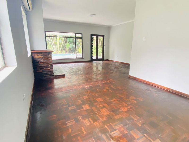 Spacious living area featuring classic parquet flooring, a brick fireplace, and large windows providing natural light and views of the garden and pool area.