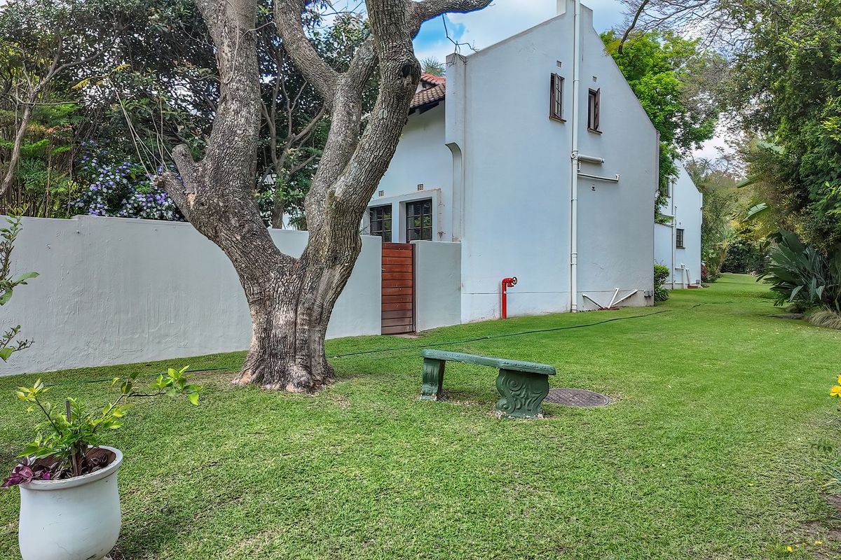 A view of the rear garden area featuring a well-maintained lawn, mature trees, and the side elevation of the property with a boundary wall.